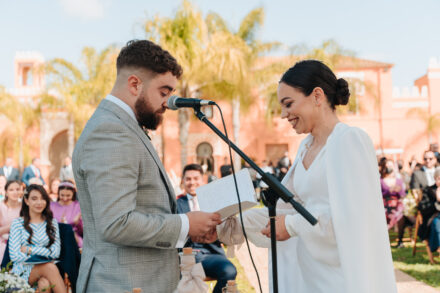 The bride smiles as the groom reads the vows before the seated guests, with the pink facade of the hacienda behind