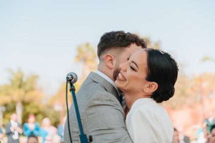 The groom hugs the bride, who laughs with her eyes closed by the ceremony microphone, at Hacienda Al-Baraka (Seville)
