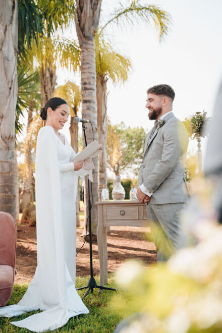The bride, in a white cape, smiles as she reads her vows to the groom beside a small table with jars of sand