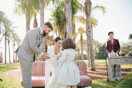 A little girl in a white dress walks up to the couple beside a pink sofa at the ceremony among palm trees