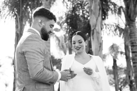 In black and white, the groom slips the ring onto the bride's finger as she smiles down at their hands