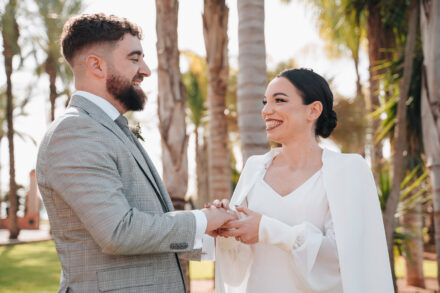 The couple smile at each other holding hands after exchanging rings, among palm tree trunks