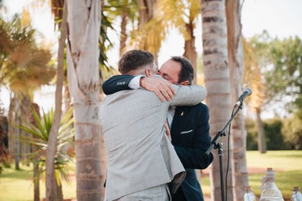 The groom shares a tight hug with a guest in a navy suit next to the ceremony microphone