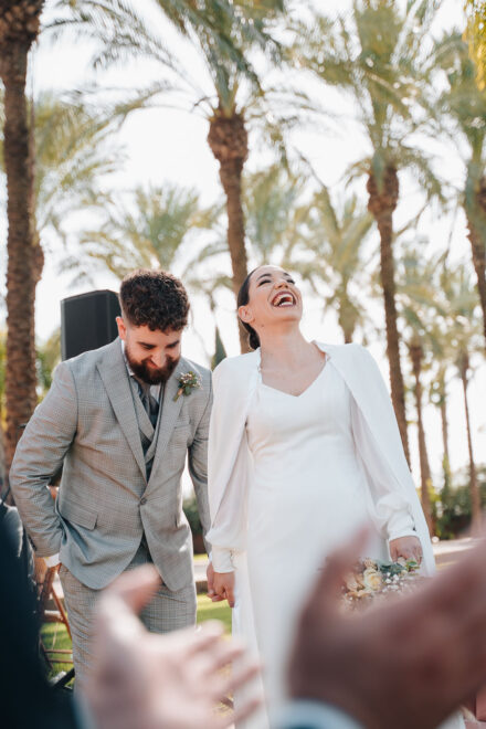 The bride bursts out laughing and the groom lowers his head with a grin as the ceremony ends, under palm trees