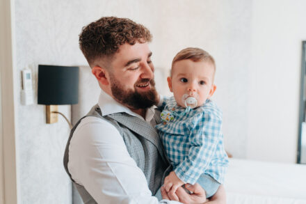 The groom smiles as he holds a baby in a blue gingham shirt with a dummy