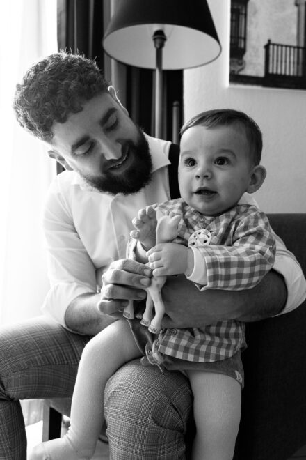 The groom sits the baby on his knee and smiles down at him, in black and white