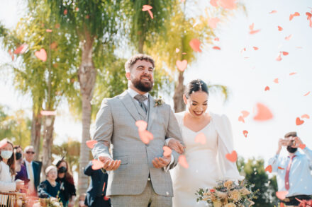 The couple smile under a shower of red heart-shaped confetti as they leave the ceremony