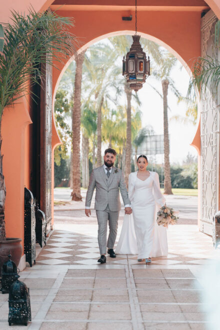 The couple walk hand in hand under a horseshoe arch with a hanging lantern and palm trees behind