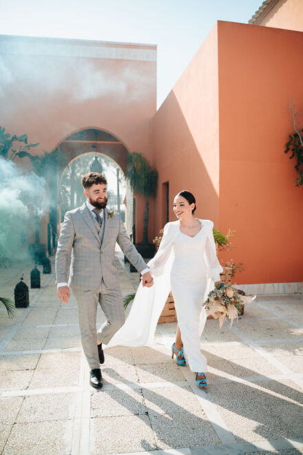 The couple walk hand in hand across the courtyard beside green smoke, her in blue shoes, at Hacienda Al-Baraka (Seville)