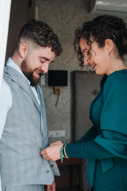 A woman in green buttons up the groom’s waistcoat as they both laugh