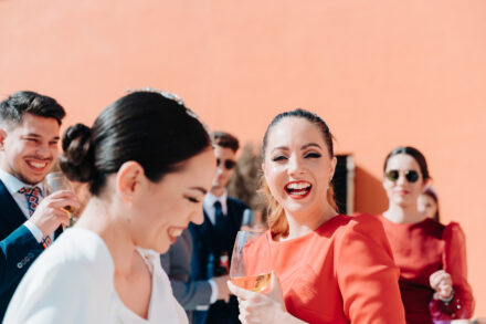The bride laughs next to a guest in orange holding a glass of wine at the cocktail hour, against an orange wall
