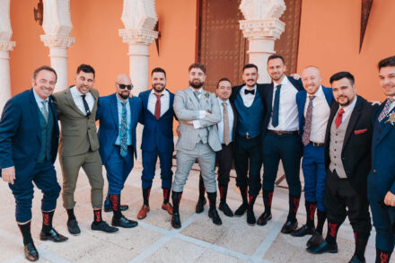 The groom poses with a group of guests, all in black socks with red lettering, among Moorish-style columns