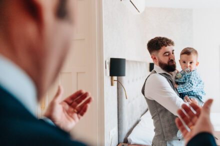 A man in a blue suit claps, out of focus, as the groom holds the baby in the hotel room