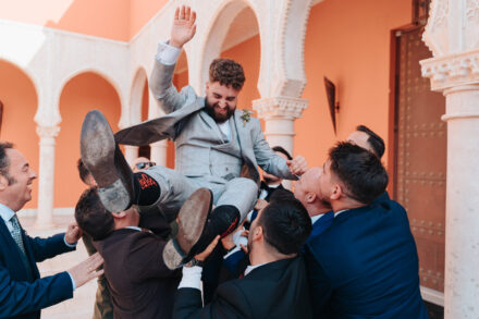 Guests toss the groom into the air as he raises a hand laughing, under the Moorish-style arches of the courtyard