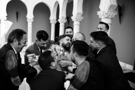 In black and white, guests hold the groom up in their arms, roaring with laughter under the courtyard arches
