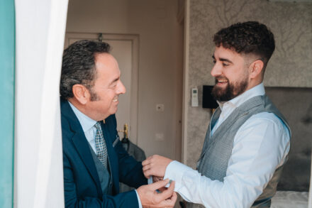 A man in a blue suit fastens the groom’s cufflinks as they smile at each other