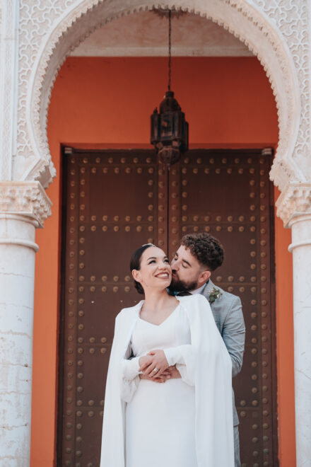 The groom hugs the bride from behind and kisses her cheek before a studded door under a carved arch