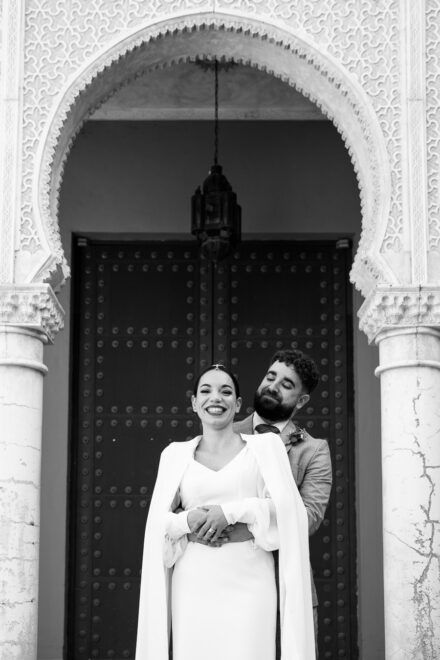 In black and white, the groom hugs the bride from behind as she laughs at the camera under the carved arch