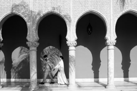 In black and white, the groom leads the bride by the hand between the arches with palm shadows