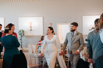 The couple cross the banquet hall hand in hand between the tables, smiling at the guests