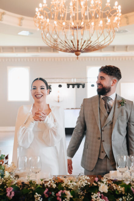 The couple laugh standing behind the flower-lined head table, under a large golden chandelier