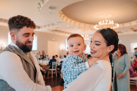 The groom looks at the baby the bride holds in her arms in the banquet hall