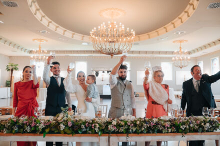 The couple, the bride holding a baby, stand and toast with the rest of the flower-decked head table