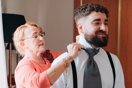 A woman in glasses and a coral dress adjusts the groom’s shirt collar and grey tie