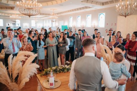Guests watch and film the couple, seen from behind with the baby, in the arched-window hall of Hacienda Al-Baraka