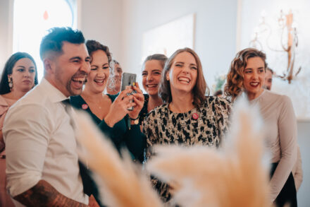 Guests burst out laughing as one records on her phone, behind out-of-focus pampas grass