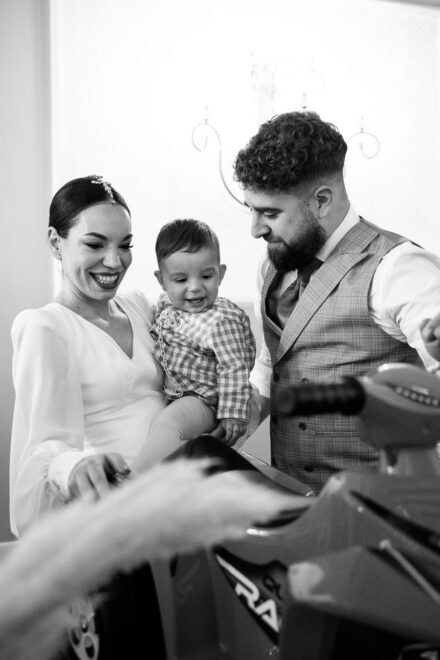 The bride and the baby smile beside the groom, who looks down at a children’s ride-on motorbike, in black and white