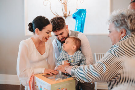 The bride and a guest open a gift box for the baby, held by the groom, next to a blue number 1 balloon