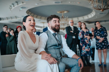 The couple laugh out loud on a white sofa while guests stand and watch