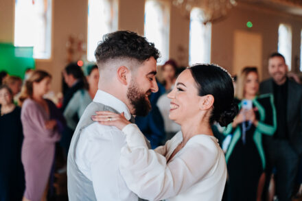 The couple smile at each other, embracing on the dance floor surrounded by guests