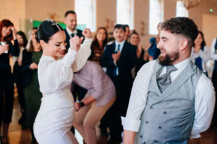 The bride dances with raised fists facing the laughing groom, as a guest crouches behind them