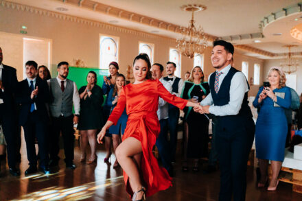 A guest in a red dress dances hand in hand with a guest in a waistcoat as the others clap