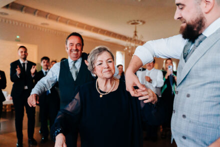 The groom dances with an older guest wearing a pearl necklace as another guest smiles behind