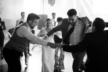 Several guests dance in a circle holding hands, with the bride smiling behind them, in black and white