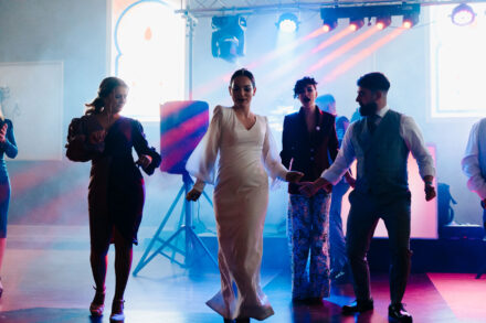 The bride dances in a line with guests amid smoke and colored stage lights during the party