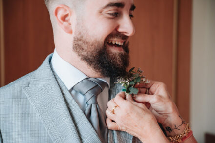 The groom laughs as a pair of hands pins a eucalyptus buttonhole to his lapel