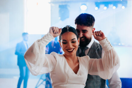 The bride dances with her eyes closed and her fists at head height, the groom laughing behind her