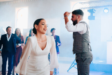 The bride dances smiling in the foreground as the groom raises his arm behind her in bluish light