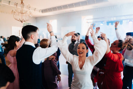 The bride dances with her arms raised, surrounded by guests, with a chandelier in the background