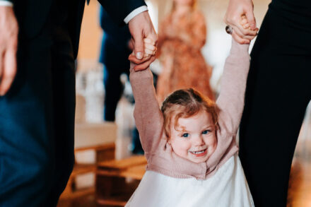 A little girl in a pink cardigan smiles as she swings from the hands of two adults during the party