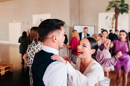 The bride laughs openly with her arm around a guest in a waistcoat during the party
