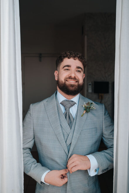 The groom, now in his grey checked suit, buttons his jacket between the curtains