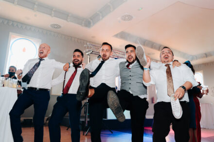 Five guests in shirts and ties kick their legs up together on the dance floor, one holding a sneaker