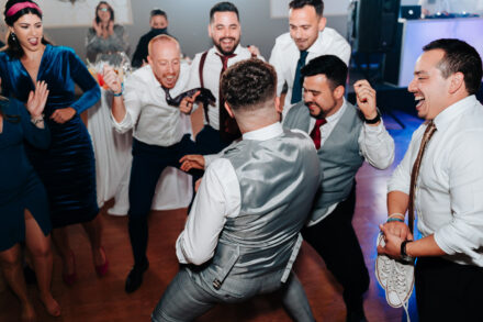 The groom, seen from behind in a gray waistcoat, dances low in a circle of laughing guests