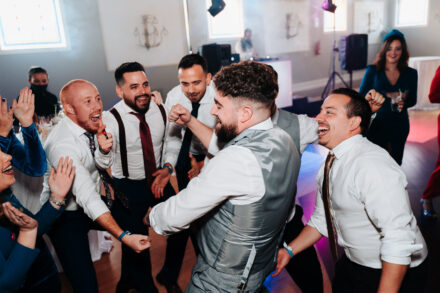 The groom dances with a raised fist, surrounded by laughing friends in ties on the dance floor