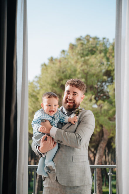 The groom and the baby laugh out loud on the balcony, with pine trees behind them